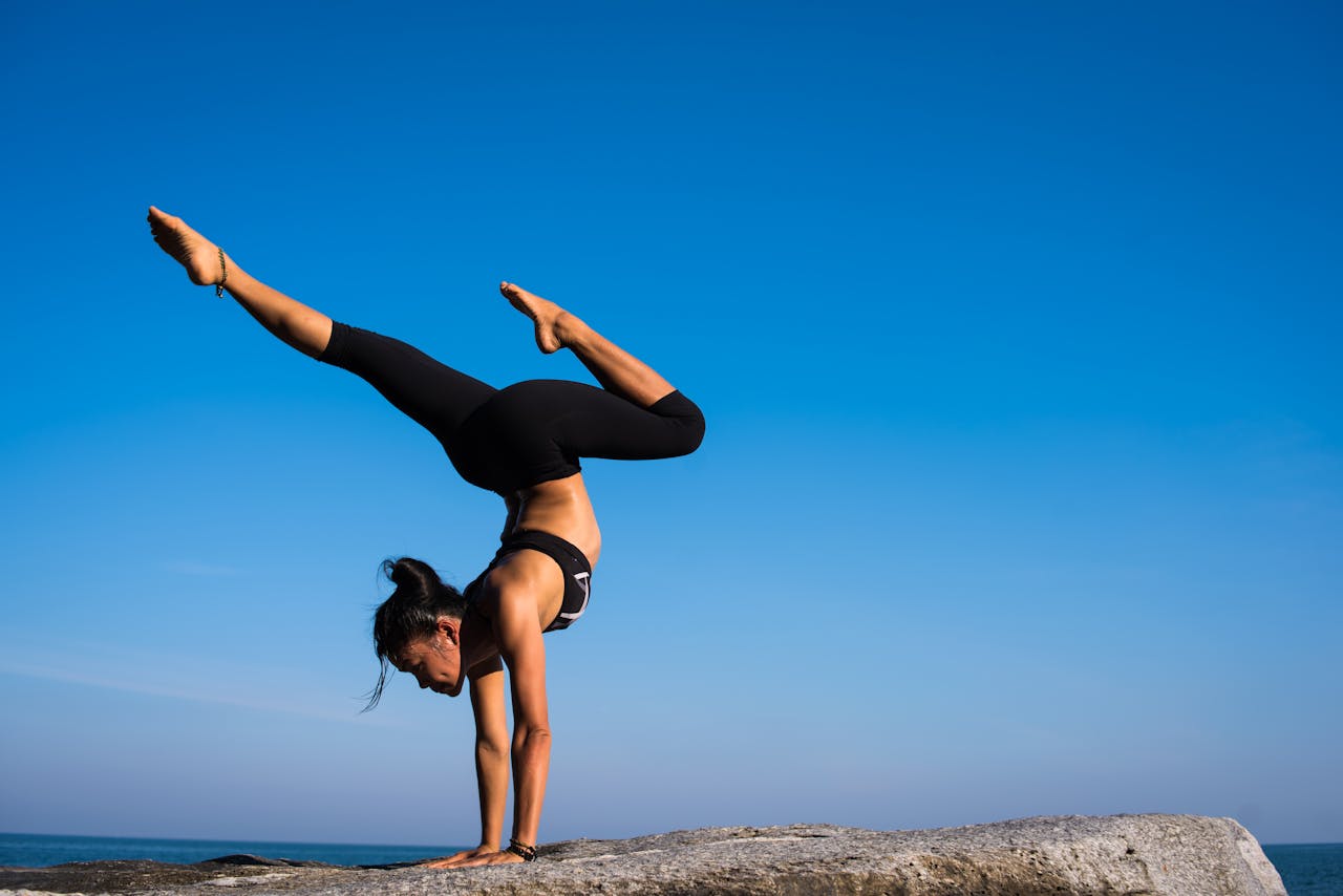 A woman performing a yoga handstand on a rock by the sea under a clear blue sky.